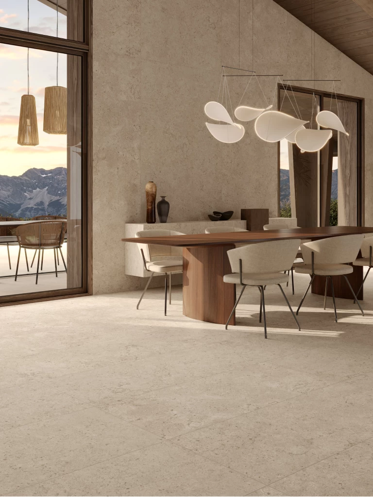 Living con zona pranzo, pavimento e pareti in gres effetto pietra Nyra Utopia Barley di Atlas Plan, abbinati a travi in legno scuro e vetrate sulle montagne.