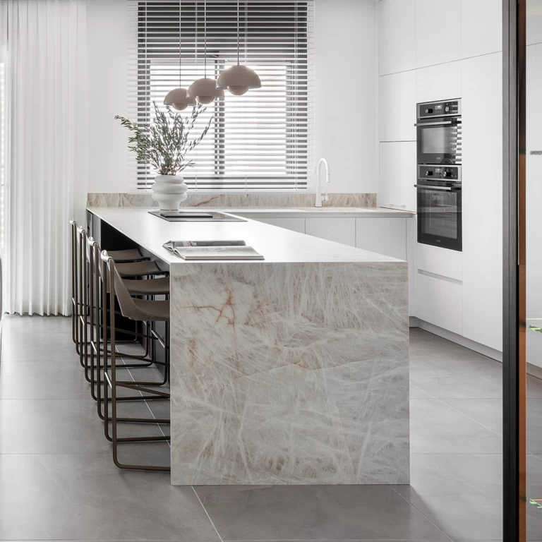 Primer plano de una cocina moderna con encimera de gres porcelánico efecto mármol Crystal White, sillas de bar de cuero marrón y un florero blanco con ramas verdes que crean un contraste delicado